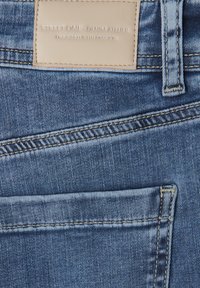 Tessuto di denim blu con superficie strutturata, che presenta una tasca posteriore visibile e un'etichetta in pelle con la scritta "STREET ONE DENIM ATELIER" in rilievo.