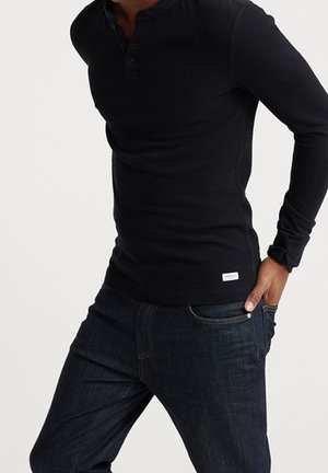 Schwarzes Langarm-Henley-Shirt aus Baumwolle mit knöpfbarem Ausschnitt und figurbetontem Schnitt. Kombiniert mit dunklen Denim-Jeans.