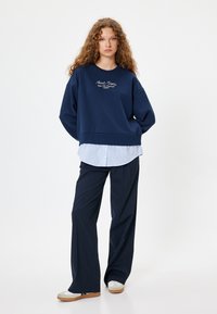 Sudadera oversized en azul marino con logo bordado, superpuesta sobre una camiseta de rayas en azul claro, combinada con pantalones marinos y zapatillas blancas.