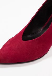 Zapato de tacón alto de ante rojo con punta afilada y diseño recortado a lo largo del costado. Interior y suela negros. Textura suave.