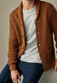 Brązowy, teksturowany kardigan z kołnierzem szalowym, zapięciami na guziki i kieszenią z przodu, noszony na szarej koszulce, zestawiony z ciemnymi dżinsami.
