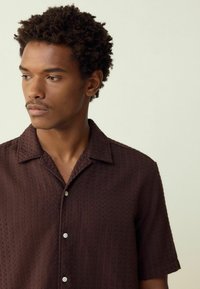 Short-sleeve, textured brown shirt with a collar and white buttons. Fabric features a subtle pattern, providing depth and visual interest.