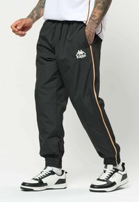 Homme portant un pantalon de survêtement Kappa noir avec des bandes latérales orange et des baskets blanches avec des accents noirs, debout avec une main dans la poche.