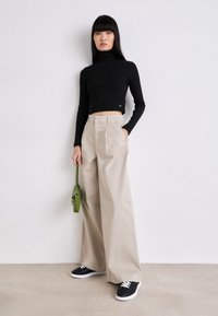 Black cropped turtleneck top, beige wide-leg trousers, black sneakers with white soles, and a small green bag. Smooth fabric, casual style.