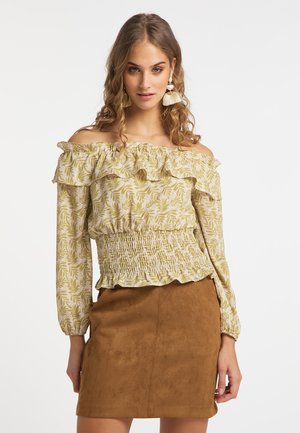 Kvinde med krøllet hår iført off-shoulder bluse med bladmønster og brun ruskindnederdel, stående mod en ensfarvet lys baggrund.