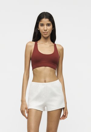 Jeune femme aux longs cheveux foncés, portant un soutien-gorge de sport bordeaux et un short blanc, debout devant un fond clair uni.