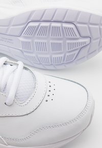 Gros plan sur une chaussure de sport blanche avec une semelle texturée, des lacets et des trous de ventilation perforés sur un fond blanc.