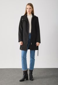 Svart, lang jakke med glidelås foran, sidelommer og krage. Båret over en lys skjorte og kombinert med blå jeans og svarte hælboots.