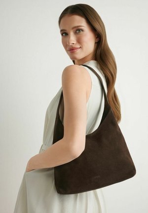 Młoda kobieta z długimi brązowymi włosami niesie ciemnobrązową zamszową torbę na ramię, ubrana w białą, bezrękawową sukienkę na jednolitym tle.