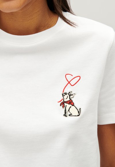 T-shirt en coton blanc avec un petit design brodé d'un chien portant une écharpe rouge et une ligne en forme de cœur rouge au-dessus.