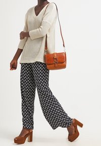 Béžový pletený svetr s výstřihem do V, kombinovaný s černými širokými kalhotami s vzory a hnědými platformními podpatky. Hnědá kožená kabelka s texturovaným detailem.