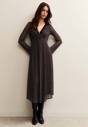 Kobieta w czarnej sukience w groszki z długim rękawem i koronkowym dekoltem, czarne botki, stojąca na tle gładkiej beżowej ściany.