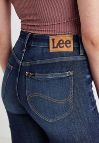 Donkerblauwe denim jeans met een hoge taille, met een bruine leren label met het "Lee" logo en gele stiksels op de zakken.