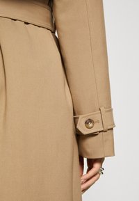 Manche de trench-coat beige avec détail de sangle boutonnée, portée par une personne avec des bagues aux doigts sur un fond uni.