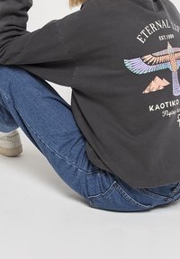 Sudadera gris de gran tamaño con un diseño gráfico que presenta un pájaro y montañas. Combinada con jeans de mezclilla azules y zapatillas blancas.