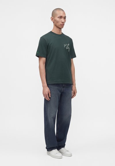 Homme debout portant un T-shirt vert foncé avec un texte blanc et un jean large délavé avec des baskets blanches sur un fond clair uni.