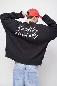 Crni pleteni džemper s spuštenim ramenima, rebrastim rubom i bijelim natpisom "Kaotiko Society" na leđima. Uparen s svijetloplavim trapericama.