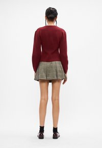 Μπορντό πλεκτή ζακέτα με μακριά μανίκια, συνδυασμένη με πτυχωτή καρό μίνι φούστα σε αποχρώσεις πράσινου, καφέ και κρέμας; φοριέται με μαύρες κάλτσες και παπούτσια.