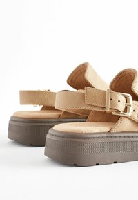 Sandales en daim beige avec une semelle plateforme, une sangle réglable à boucle et une finition lisse. La semelle présente des détails de grip texturé.
