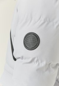 Gray puffer jacket fabric with a smooth texture, featuring a round black logo patch on the sleeve. Stitching details are visible.