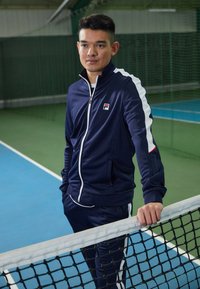 Marineblauwe trainingsoutfit met witte zijkanten, bestaande uit een ritsjack met opstaande kraag en slim-fit broeken, weergegeven tegen een achtergrond van een tennisbaan.