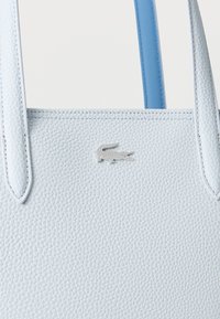 Sac à main en cuir texturé bleu clair avec des bretelles fines et un petit logo crocodile argenté centré sous les poignées.
