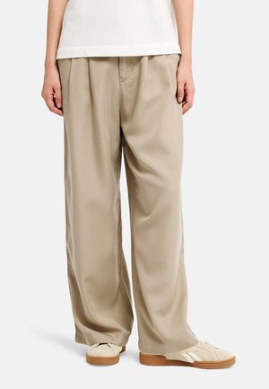 Pantaloni beige a gamba larga abbinati a sneakers bianche con suola in gomma e camicia bianca, mostrati dalla metà del busto ai piedi.