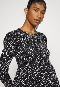 Vestido preto com mangas longas bufantes, apresentando um padrão de bolinhas brancas. Tecido suave com um corpete justo e uma cintura franzida.