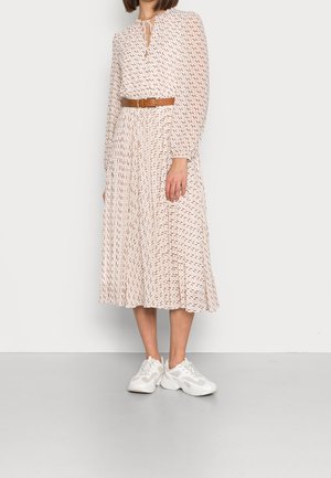 Frau trägt ein hell gemustertes Midikleid mit langen Ärmeln, einem braunen Gürtel und weißen klobigen Sneakers und steht vor einem schlichten Hintergrund.