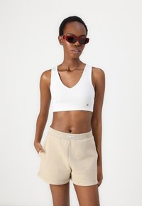 Bijeli rebrasti crop top s V-izrezom i malim logotipom, u kombinaciji s bež kratkim hlačama na elastik, koje imaju bočne džepove. Tamne pravokutne sunčane naočale.