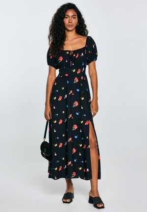 Zwart bloemenmaxi-jurk met korte pofmouwen, voorzien van kleurrijke bloempatronen, een strik bij de halslijn en een zijsplit. Geaccentueerd met een handtas.