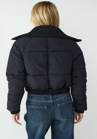 Czarna krótka kurtka puchowa z prążkowanym kołnierzem, pikowanym wzorem i elastycznym pasem, noszona na niebieskich dżinsach.