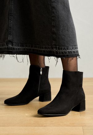 Sorte ruskind ankelstøvler med firkantet tå og blokhæl, med en sidelukning og glat tekstur. Båret med en slidset sort denimnederdel.