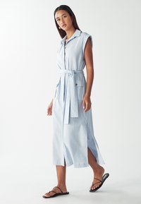 Donna in piedi che indossa un abito midi azzurro chiaro senza maniche con bottoni frontali, cintura, tasche, spacchi laterali e sandali neri con cinturini piatti.