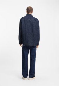 Camisa de mezclilla oscura con un ajuste relajado, mangas largas y puños con botones, combinada con pantalones sueltos de mezclilla oscura y zapatos negros.