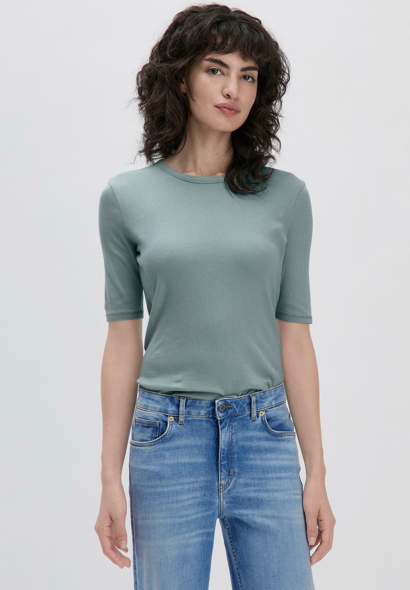 Vihreä lyhythihainen t-paita, joka on valmistettu pehmeästä kankaasta ja jossa on pyöreä kaula-aukko. Yhdistetty vaaleansinisiin farkkuihin, joissa on taskut ja kontrastitikkaukset.