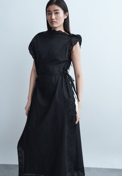 Jeune femme aux longs cheveux foncés portant une robe noire sans manches avec des bordures en dentelle, sur un fond clair uni.