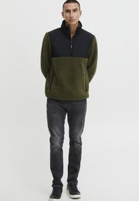 Olive green fleece pullover with a black nylon top, half-zip front, and side pockets; paired with dark jeans and black sneakers.