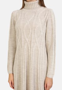 Robe pull beige en tricot avec un col roulé haut, présentant un motif en biais et des accents côtelés sur les manches et les côtés. Manches longues.