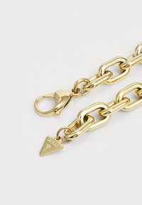 Collana in metallo dorato con maglie ovali e rettangolari intrecciate, dotata di chiusura a moschettone e un ciondolo triangolare inciso con la scritta "GUESS".