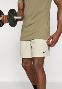 Light beige shorts and a fitted olive green t-shirt. The shorts feature a small black Nike logo. The subject is lifting a black dumbbell.