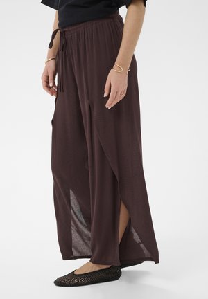 Žena oblečená v voľných tmavohnedých nohaviciach so širokými nohavicami a bočnými rozparkami, čiernych pletených balerínach, zlatých náramkoch a čiernom topu, stojaca na bielom pozadí.