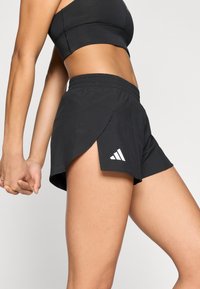 Shorts de sport noirs à coupe ample avec des fentes latérales. Dotés d'une ceinture élastique et d'un logo Adidas blanc sur le bas côté gauche.