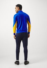 Giacca sportiva blu con dettagli gialli, abbinata a pantaloni blu navy. Texture morbida, design aderente, maniche lunghe e polsini a coste.