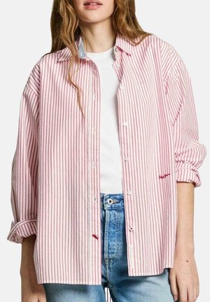 Jonge vrouw draagt een losse roze-witte verticaal gestreepte blouse over een wit topje en blauwe spijkerbroek.
