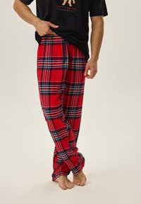 Czerwone flanelowe spodnie piżamowe w kratę z niebieskimi i żółtymi akcentami, luźny krój, elastyczna taliatura, noszone z czarną koszulką z krótkim rękawem, boso.
