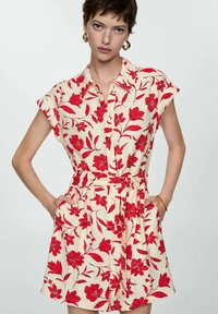 Korte mouw, beige jurk met een rood bloemenpatroon, knoopsluiting aan de voorkant, een ceintuur in de taille en zijzakken. Lichtgewicht stof met een gladde textuur.