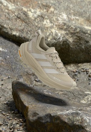 Zapatilla de senderismo Adidas Terrex de color beige claro flotando sobre terreno rocoso, mostrando la adherencia de la suela y las características del material impermeable Gore-Tex.