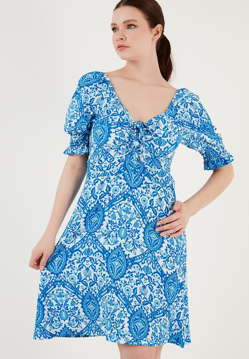Rochie cu un model alb și albastru, cu mâneci puff, decolteu în V și detaliu cu fundă în față. Fabricată dintr-un material ușor, până la genunchi.