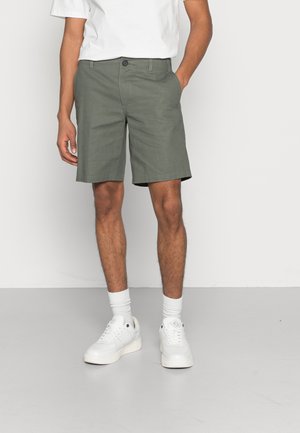 Grønne shorts med knaplukning og to frontlommer, parret med hvide sneakers og sokker. Glat materiale og lige snit.